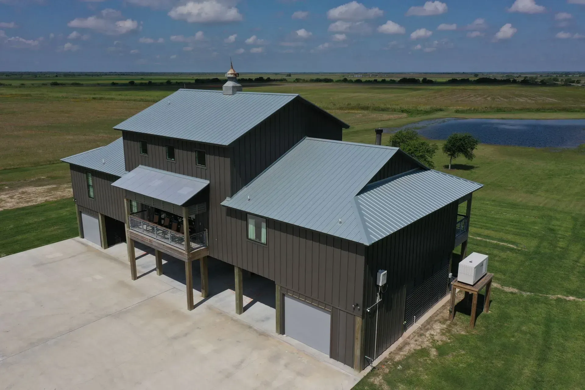 Brown barn-style building with a silver roof and multiple levels, set in a grassy field with a pond.