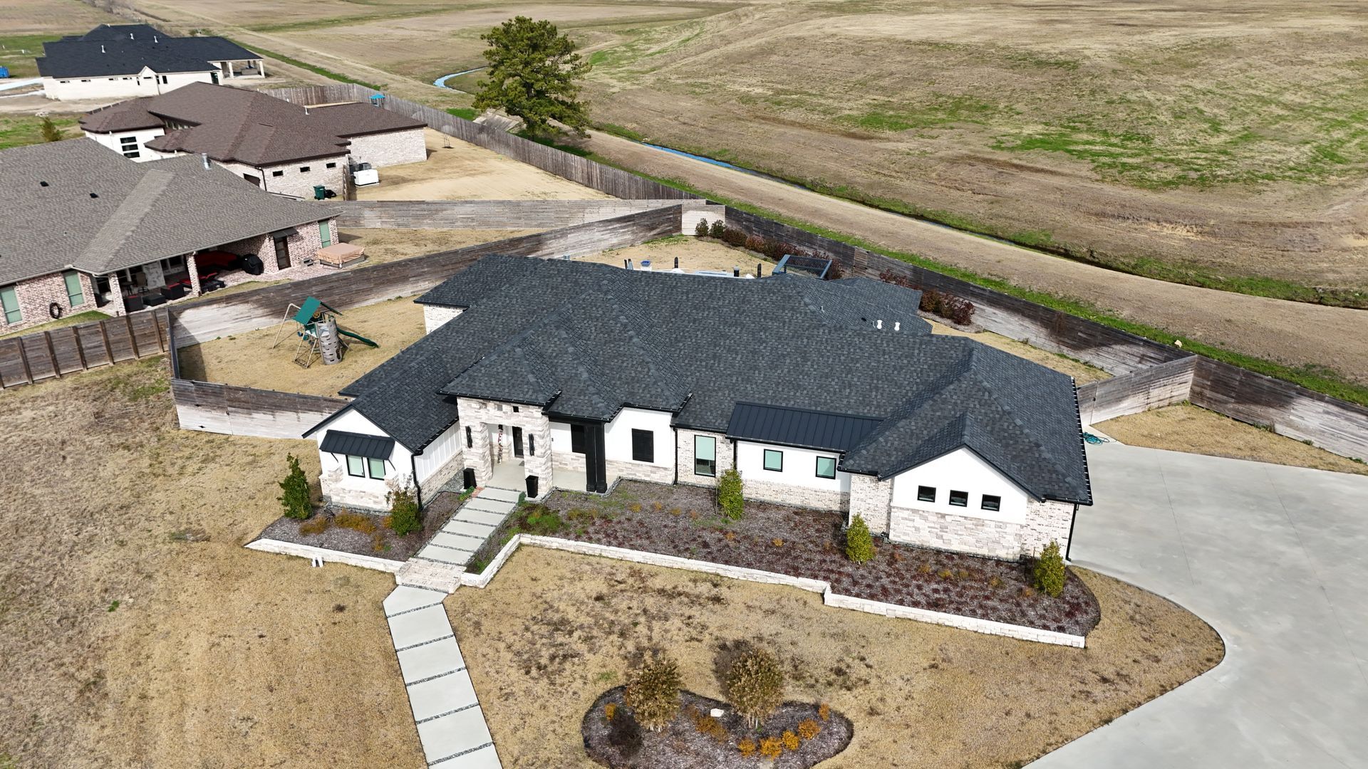 Aerial view of a modern house with a dark roof and light brick exterior; paved walkway leads to the entrance.