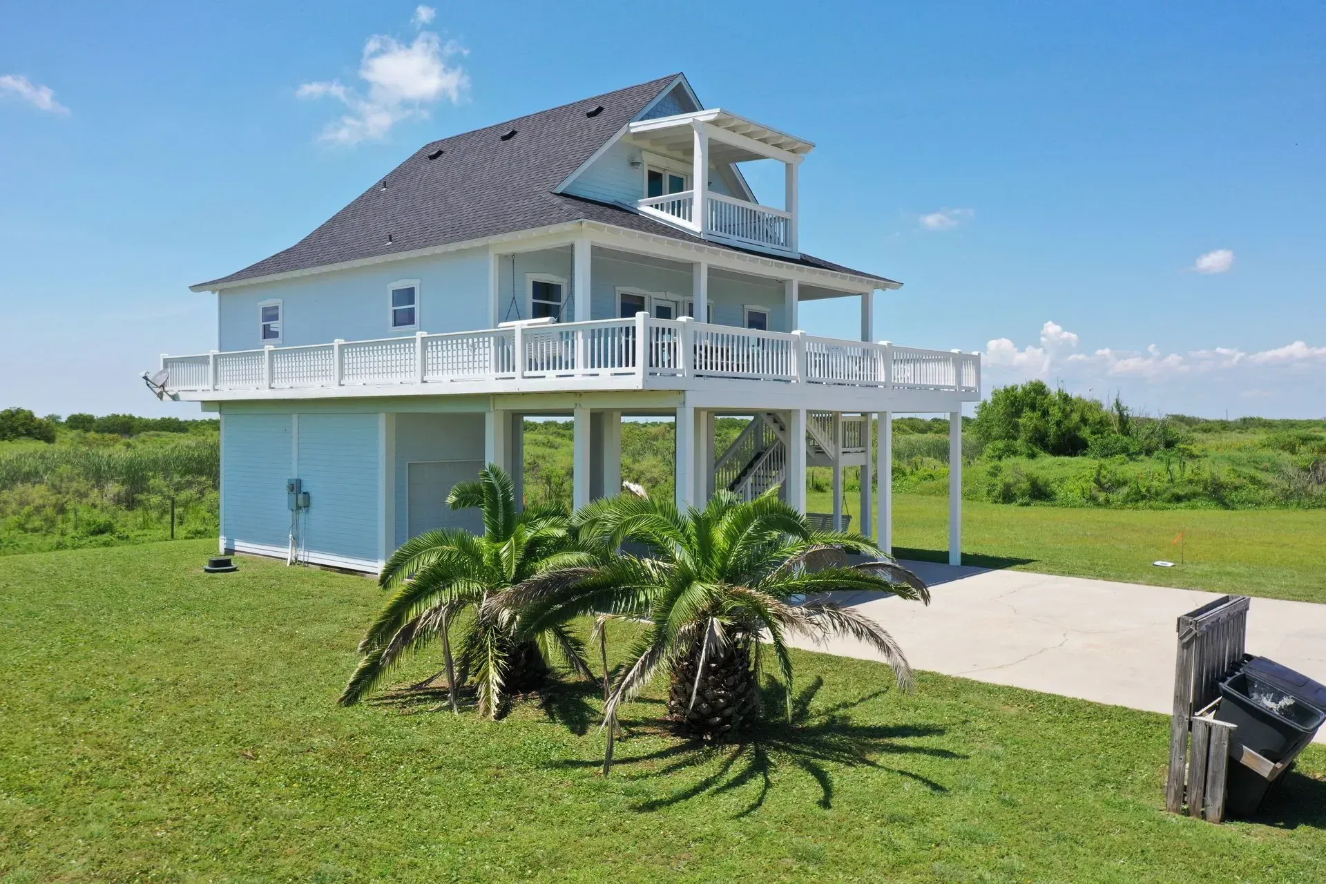 Light blue beach house on stilts with a balcony, palm trees, and green grass.