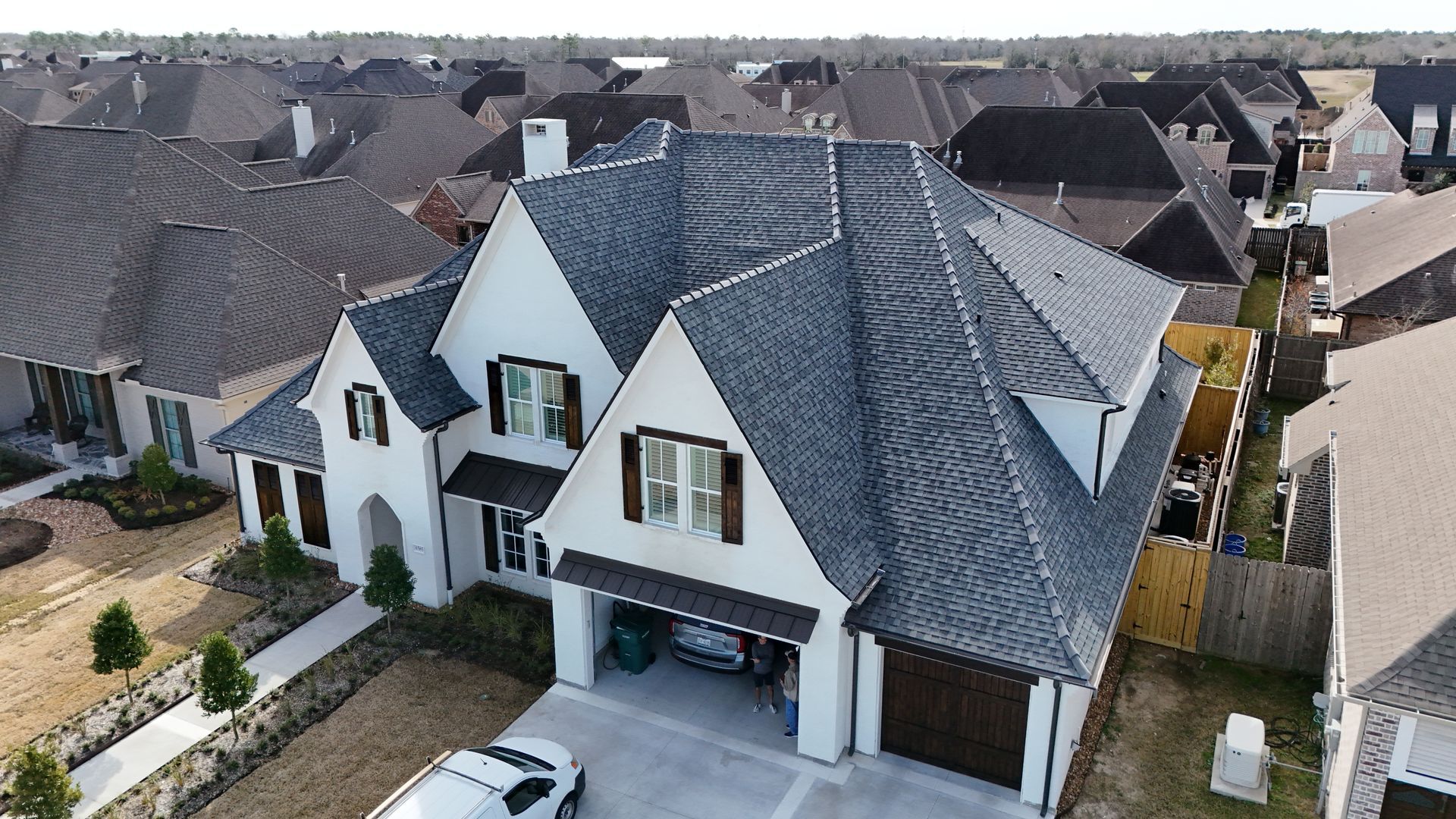 White two-story house with dark gray roof and brown garage door, surrounded by similar houses in a suburban setting.