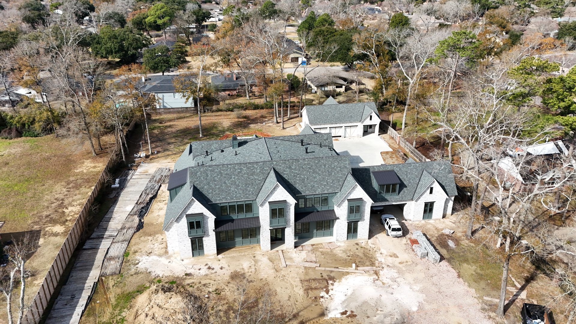 Aerial view of a large, two-story house with a detached garage. Grey roof and white exterior. Brown surroundings.