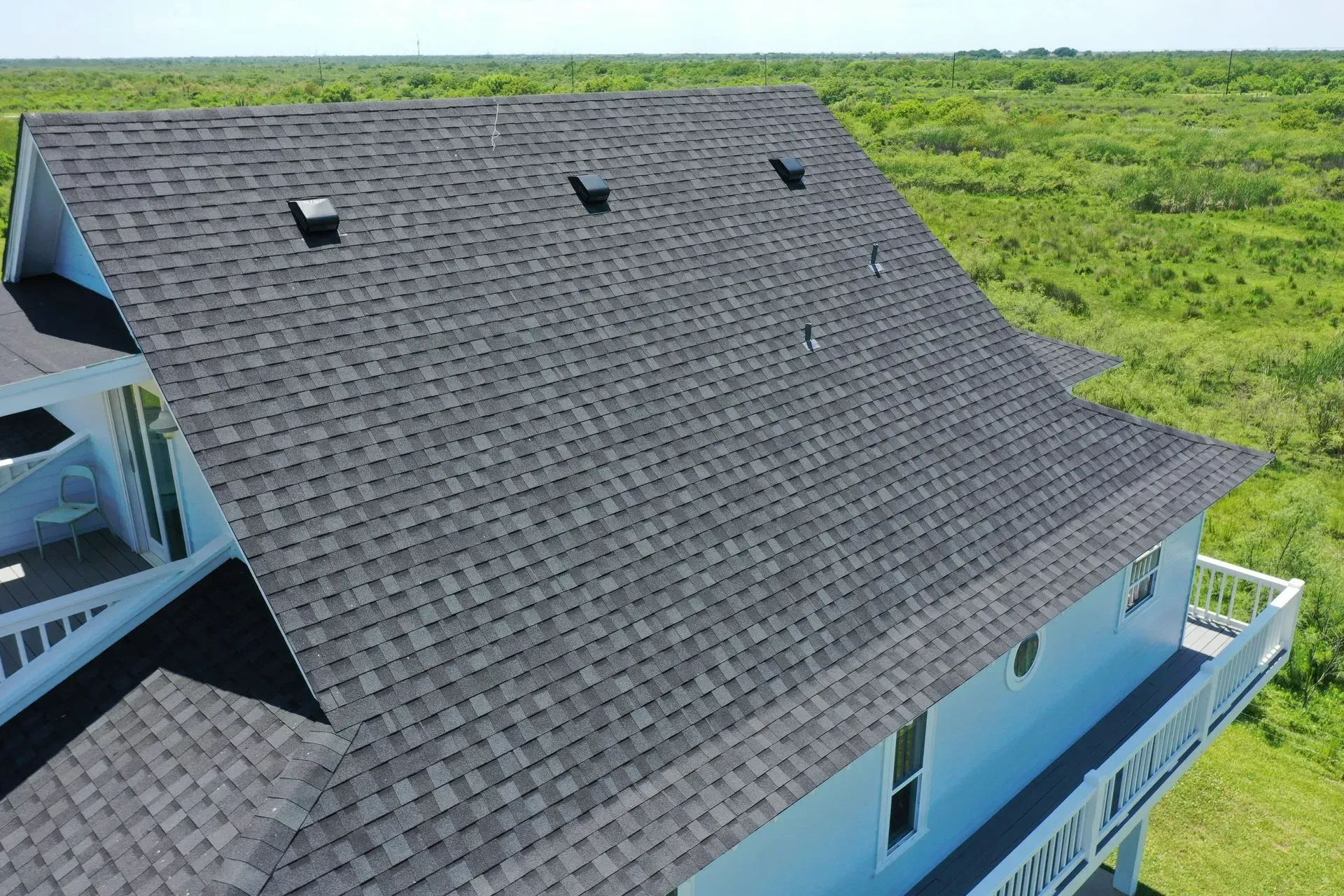 Aerial view of a light blue house with a dark gray shingle roof, overlooking a green landscape.