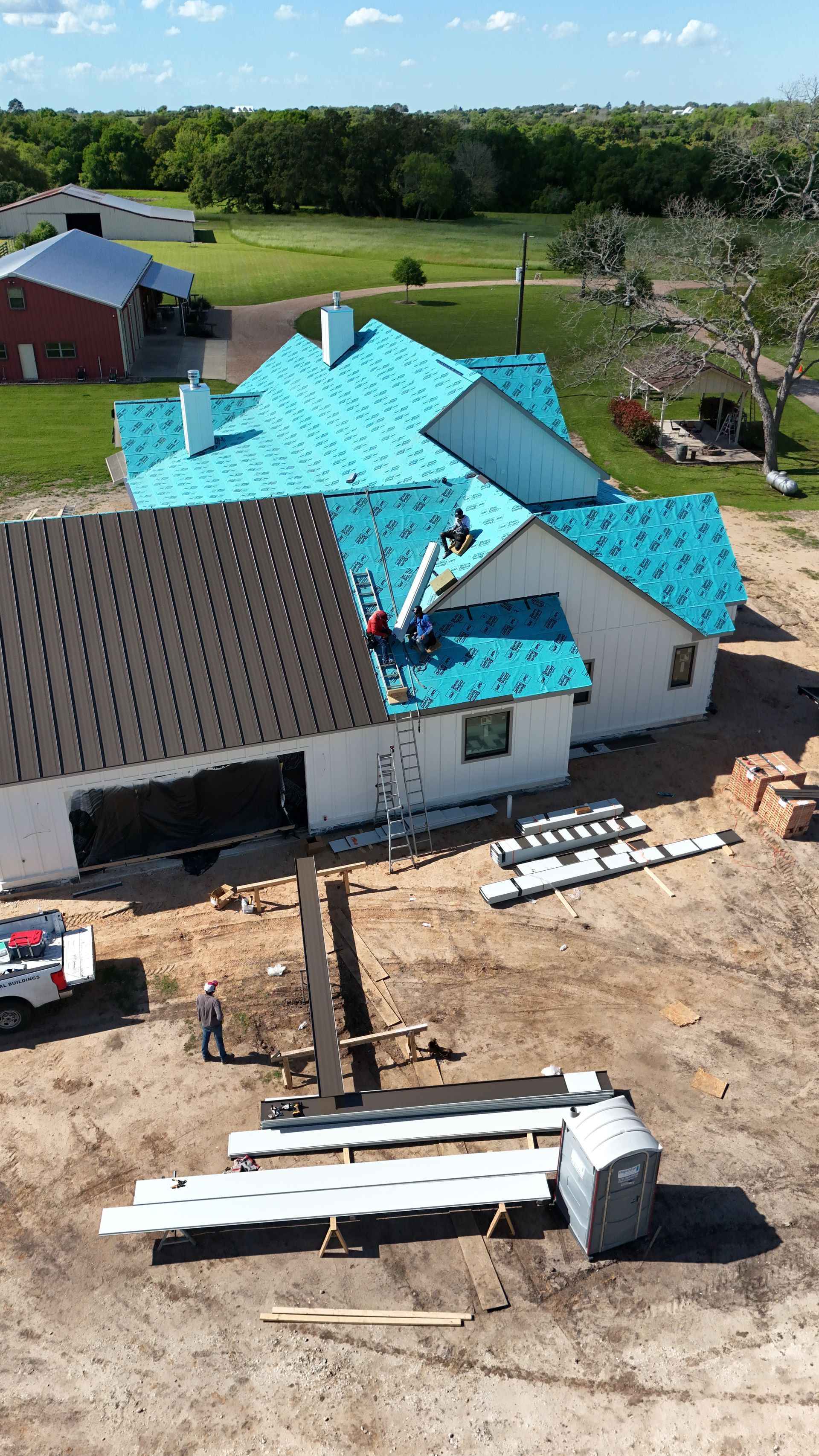 Construction of a house: workers on roof, new blue underlayment and gray shingles. Dirt ground, white house, clear day.