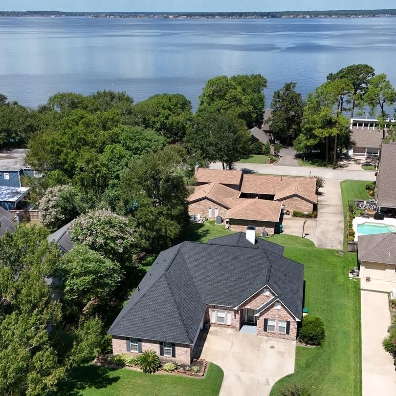 Houses with dark roofs and green lawns along a body of water with trees and sky.