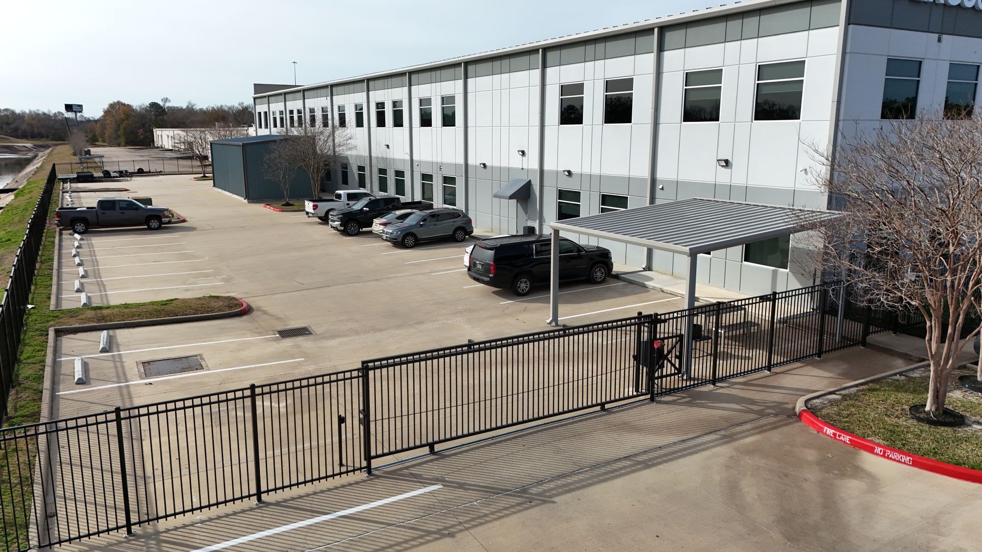 Exterior of a modern industrial building with a gated entrance and parked cars in the lot.
