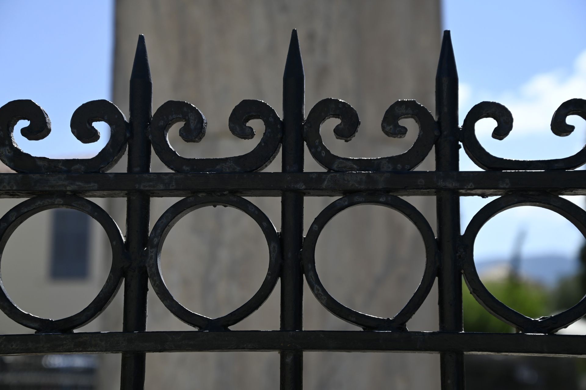 Black wrought-iron fence with decorative swirls and spikes, in front of a blurred light-colored building and sky.