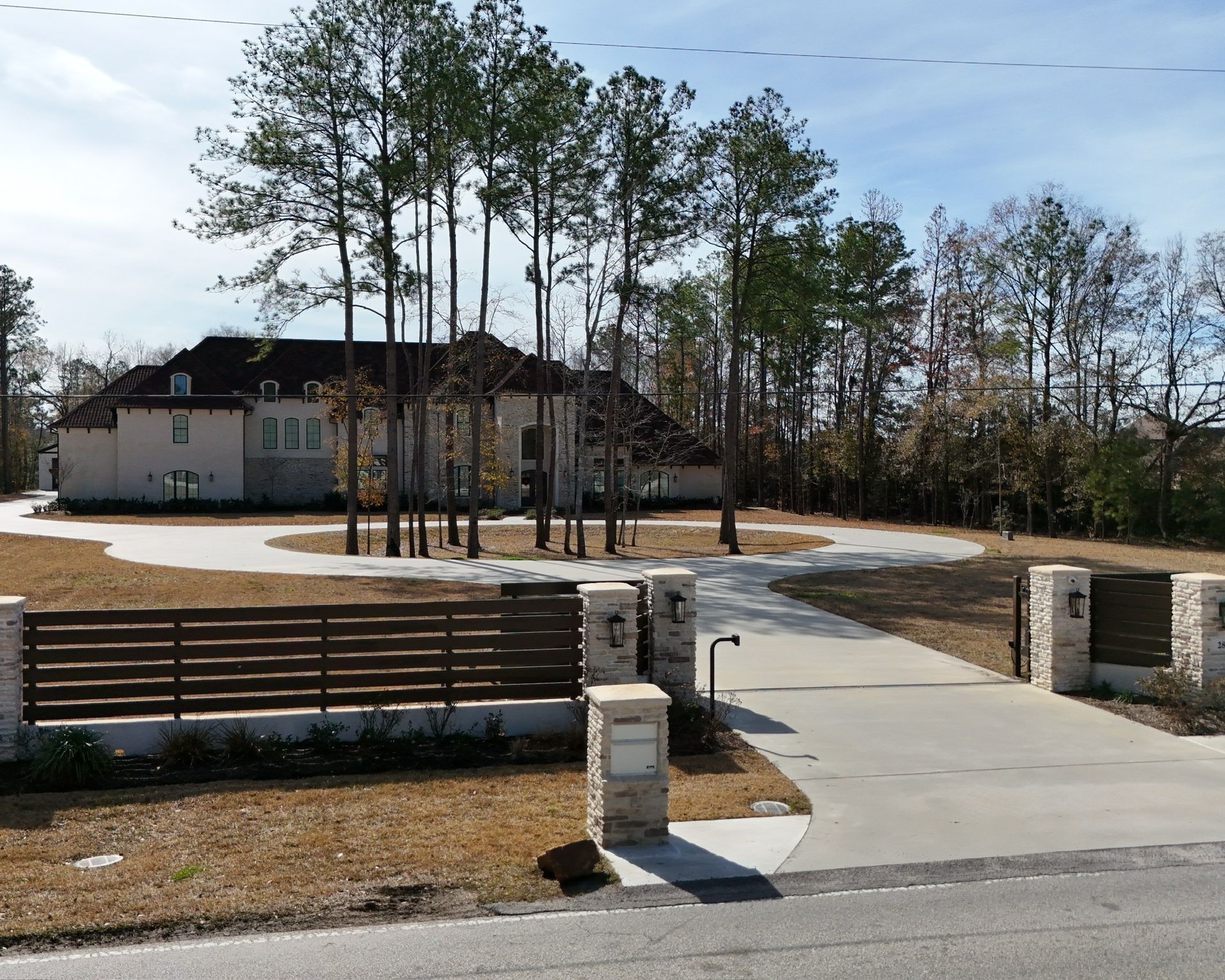A large home with a circular driveway and stone and wood fence in front. Tall pine trees in the background.