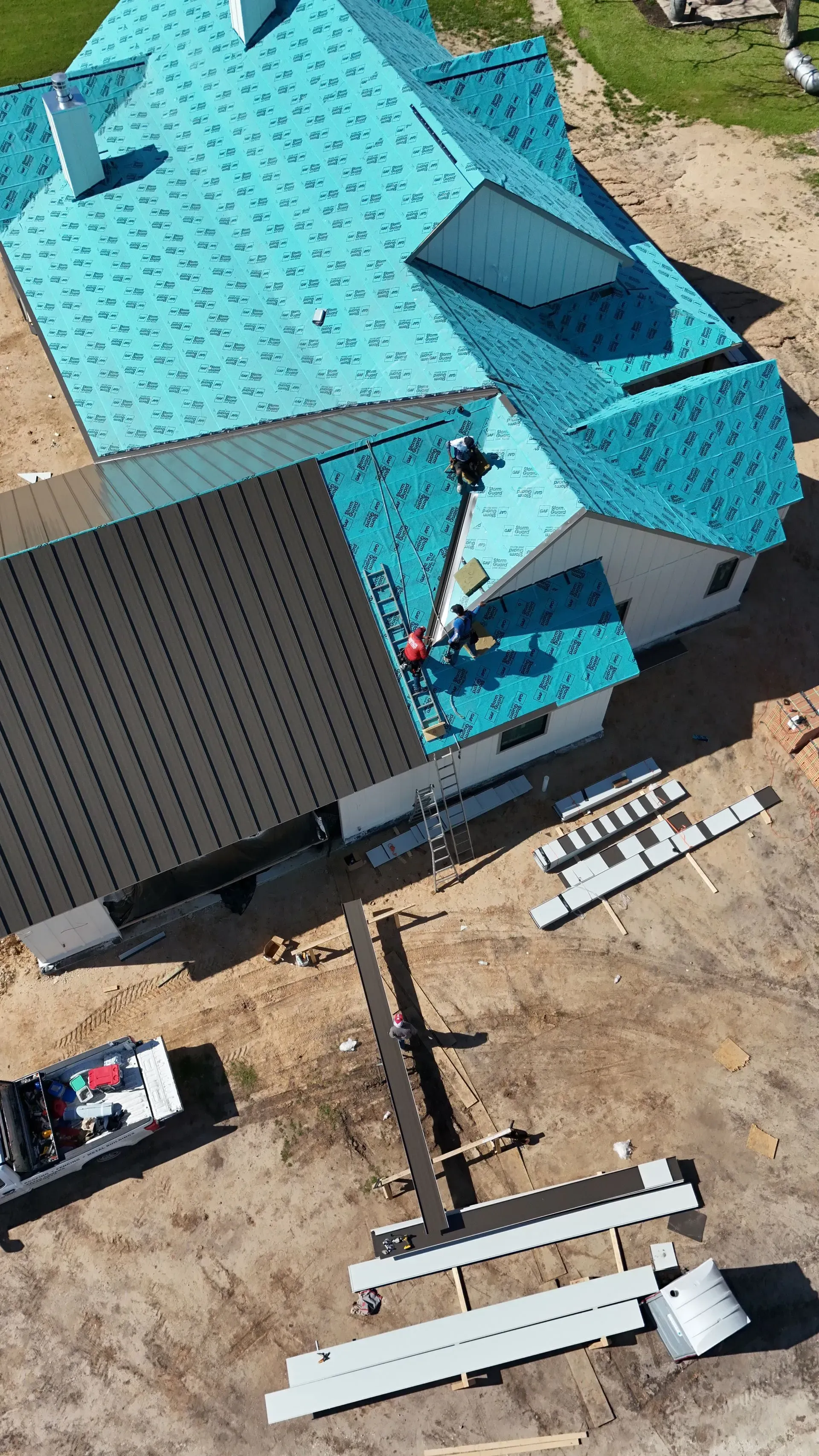 Workers installing turquoise roofing on a house, construction materials visible on the ground.