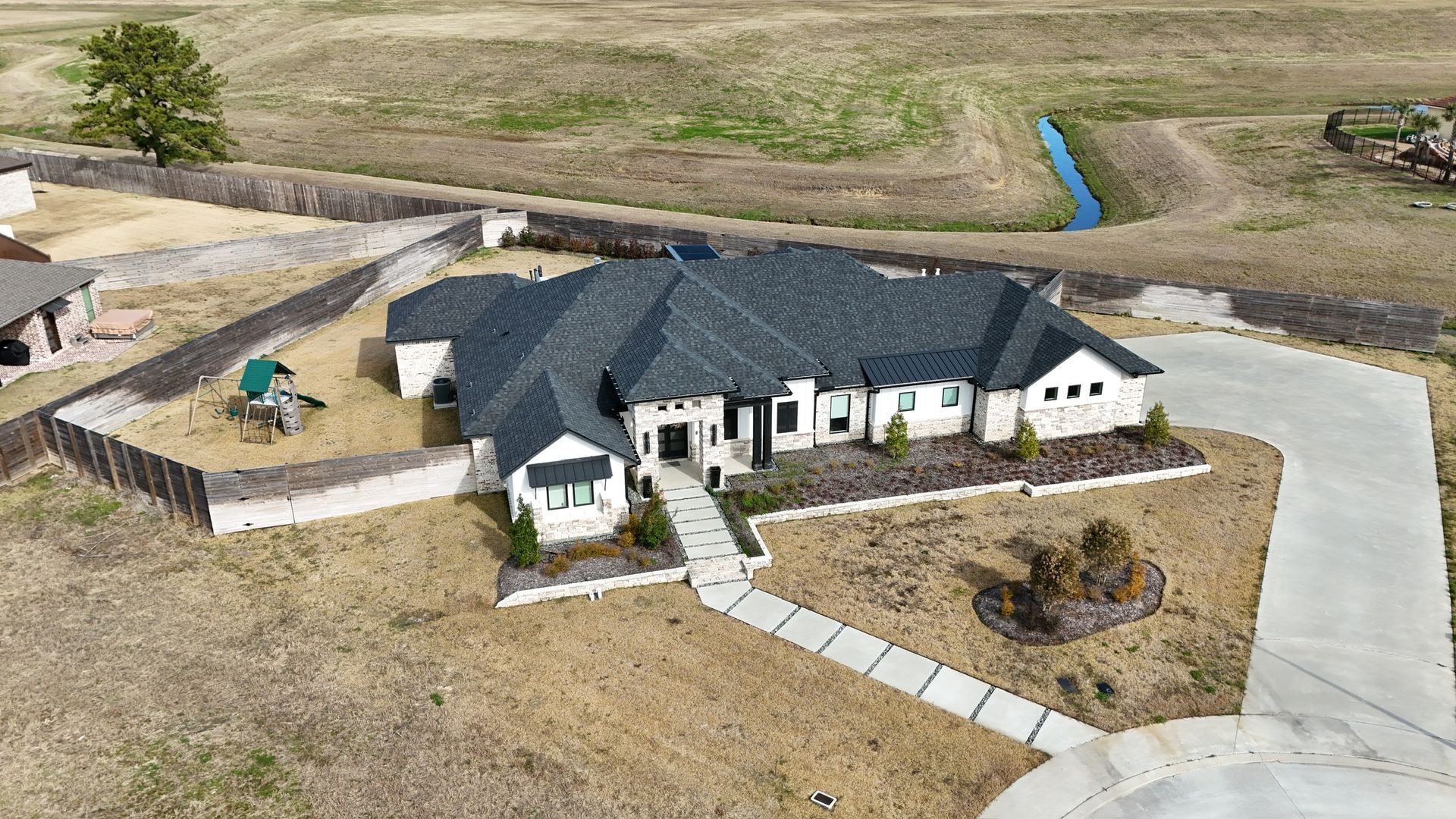 An aerial view of a single-story house with a long driveway on a brown field.