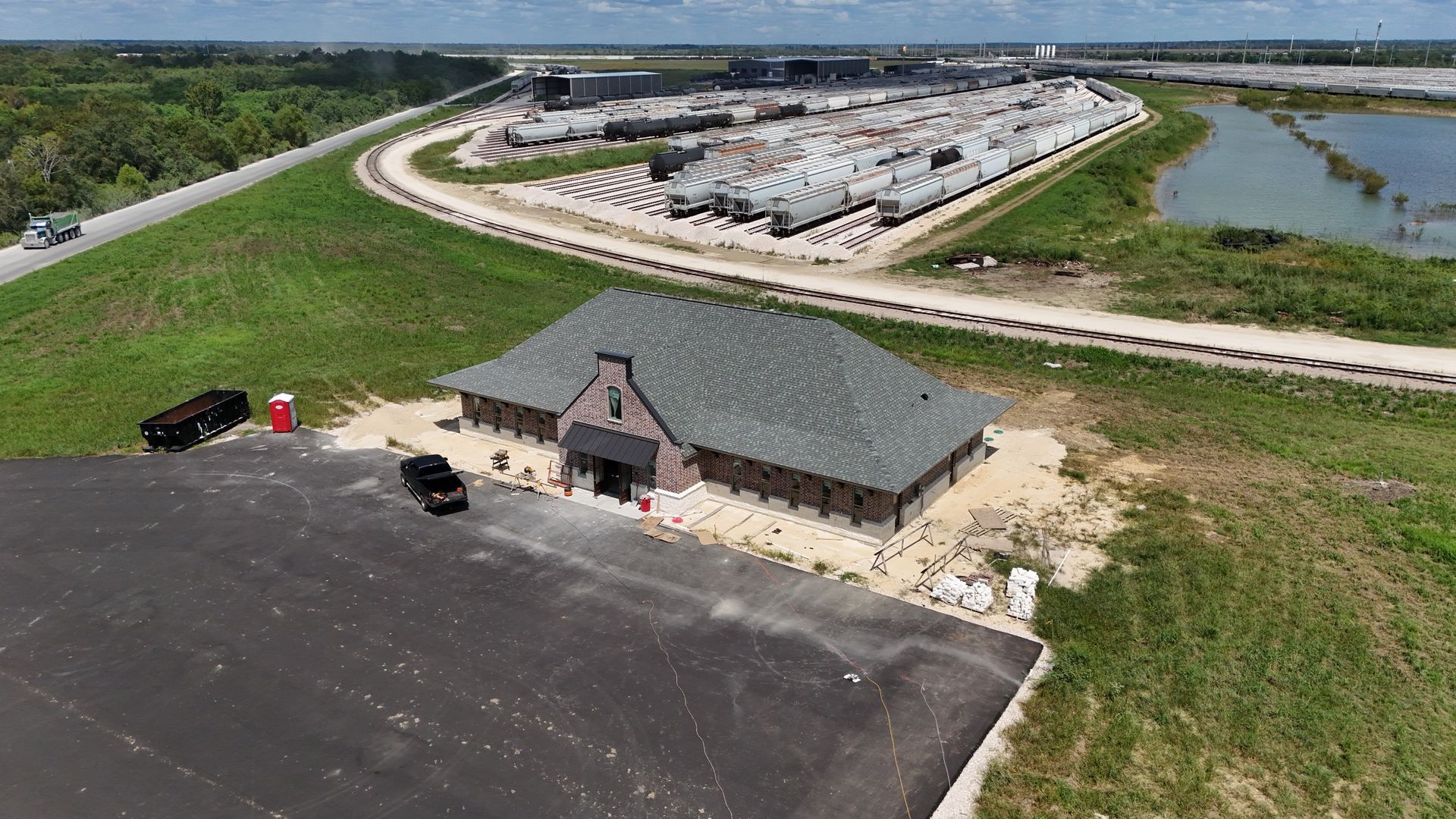 Aerial view: new building with dark roof, parking lot, nearby construction, and a road.
