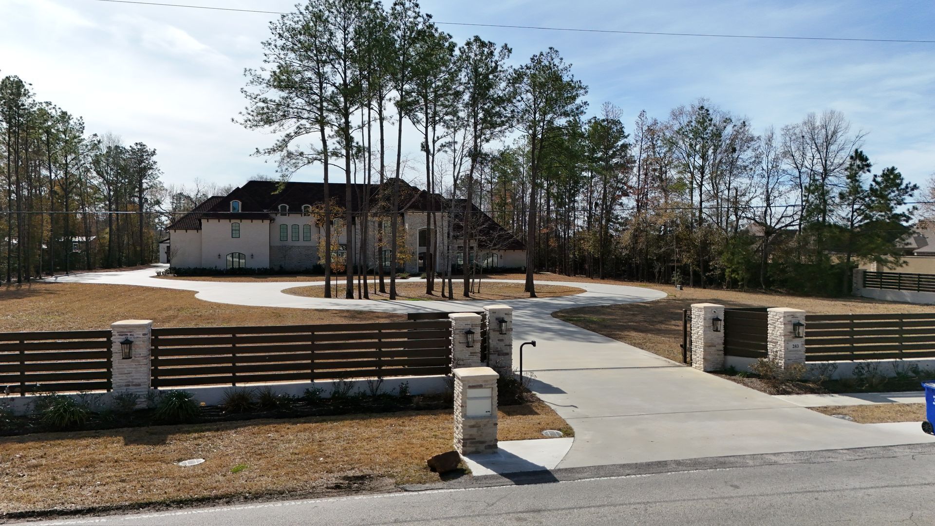 A large beige house with a long driveway and a wooden fence, on a sunny day.