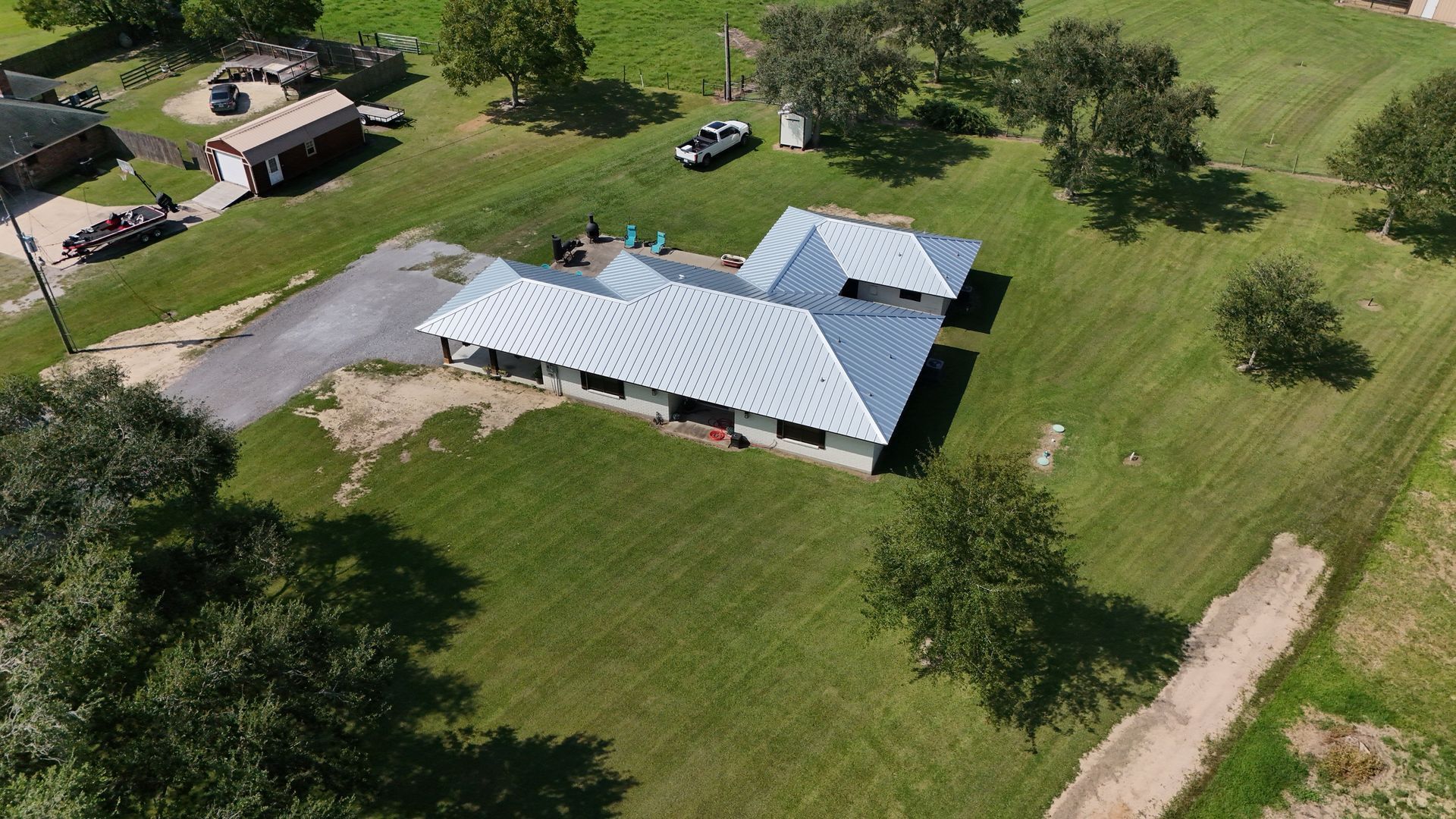 Aerial view of a white-roofed house on a grassy lot, trees, driveway, and nearby buildings.