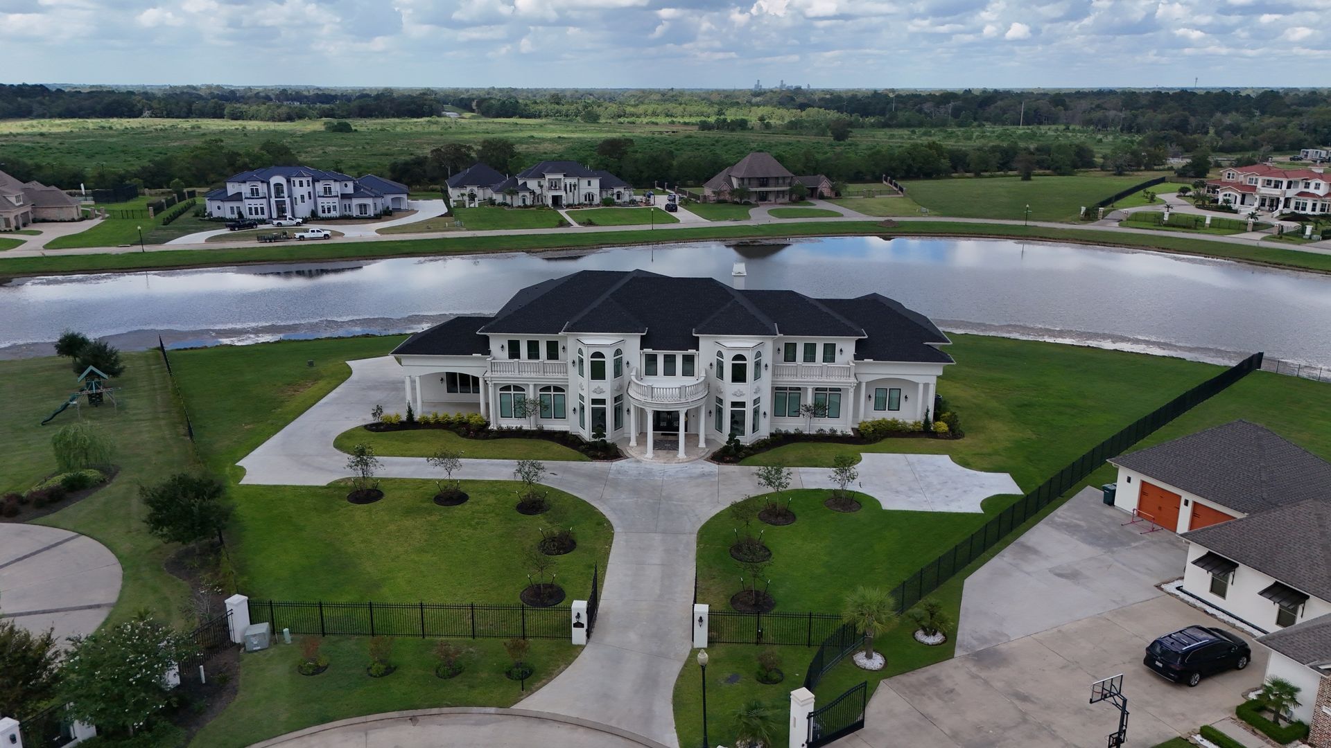 Aerial view of a large white house with a black roof, near a lake, green grass, and other houses.