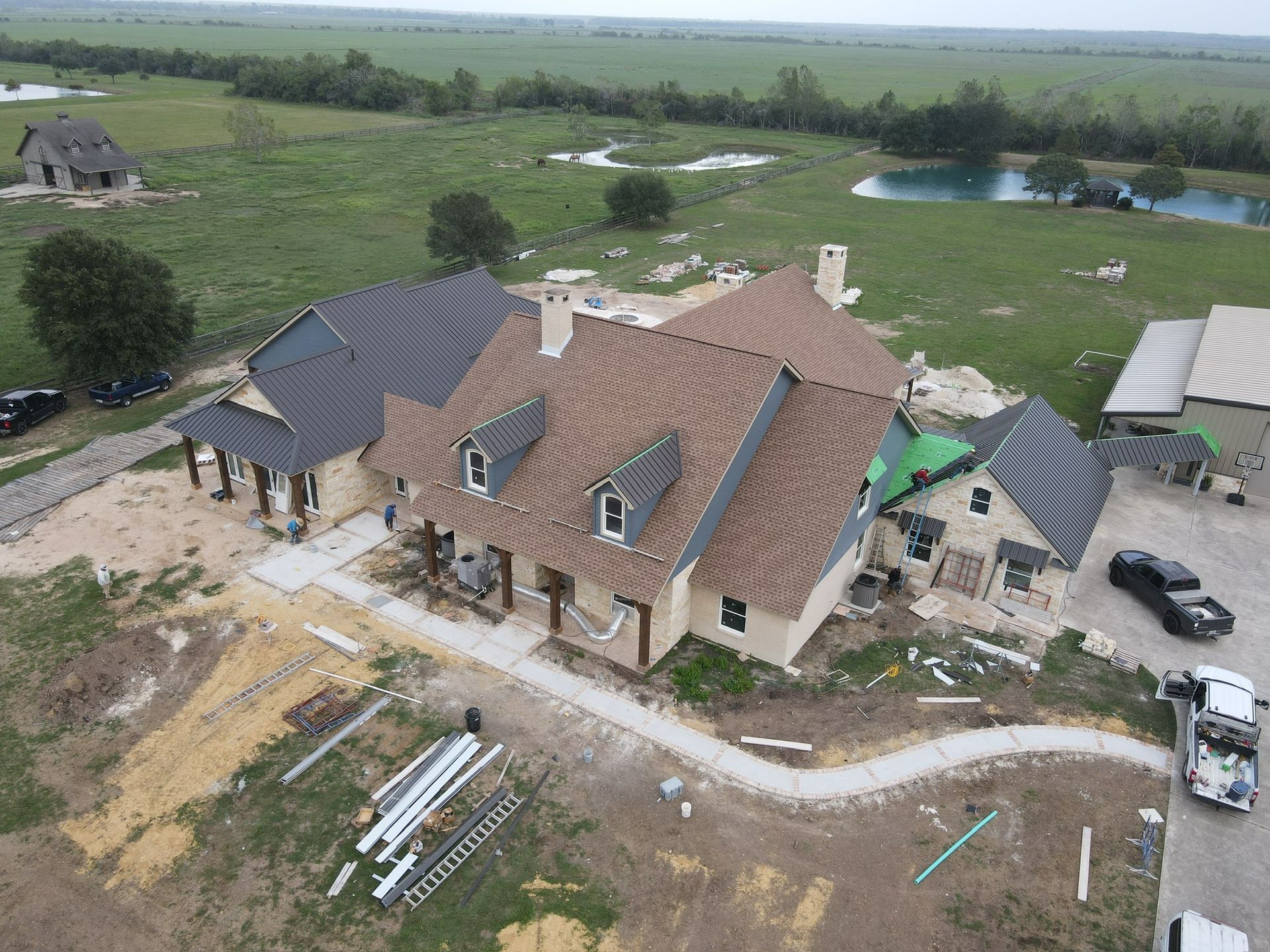 Aerial view of a house under construction with brown and gray roofs, surrounded by a grassy field and ponds.