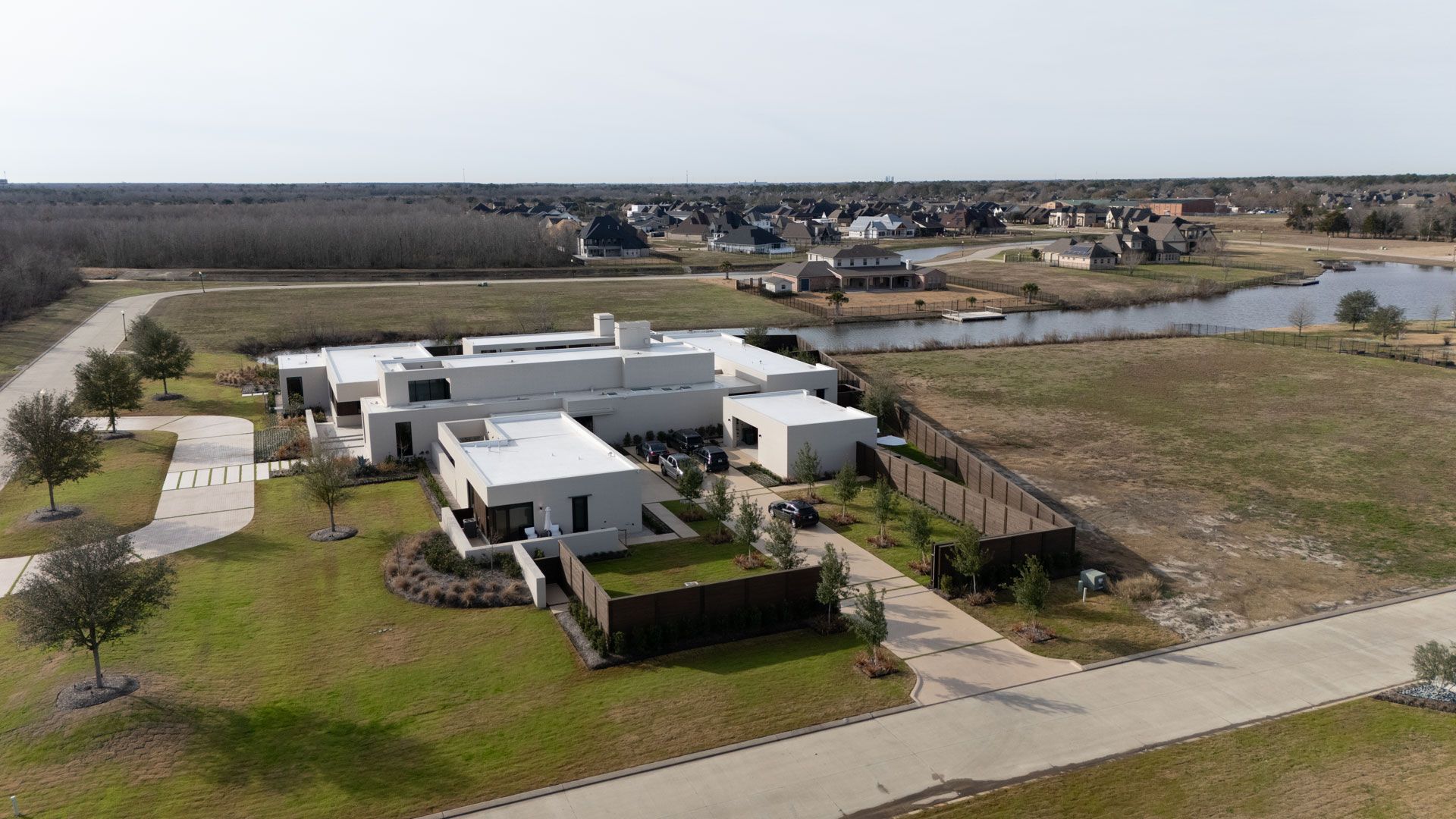 Aerial view of a modern white house with a flat roof, green lawn, and nearby pond in a suburban area.