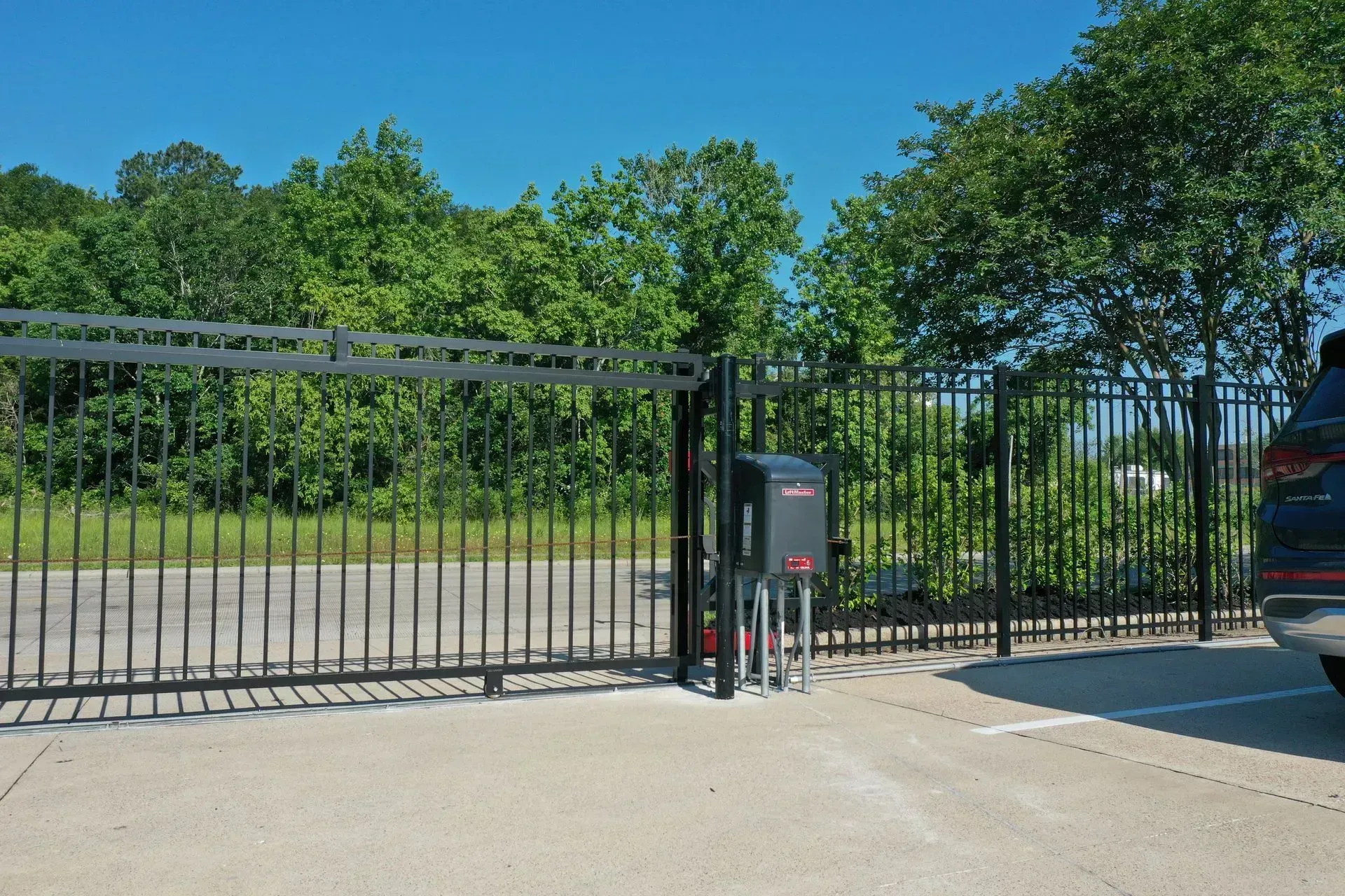 Sliding black gate with motor and a car parked in front, with trees and blue sky in the background.