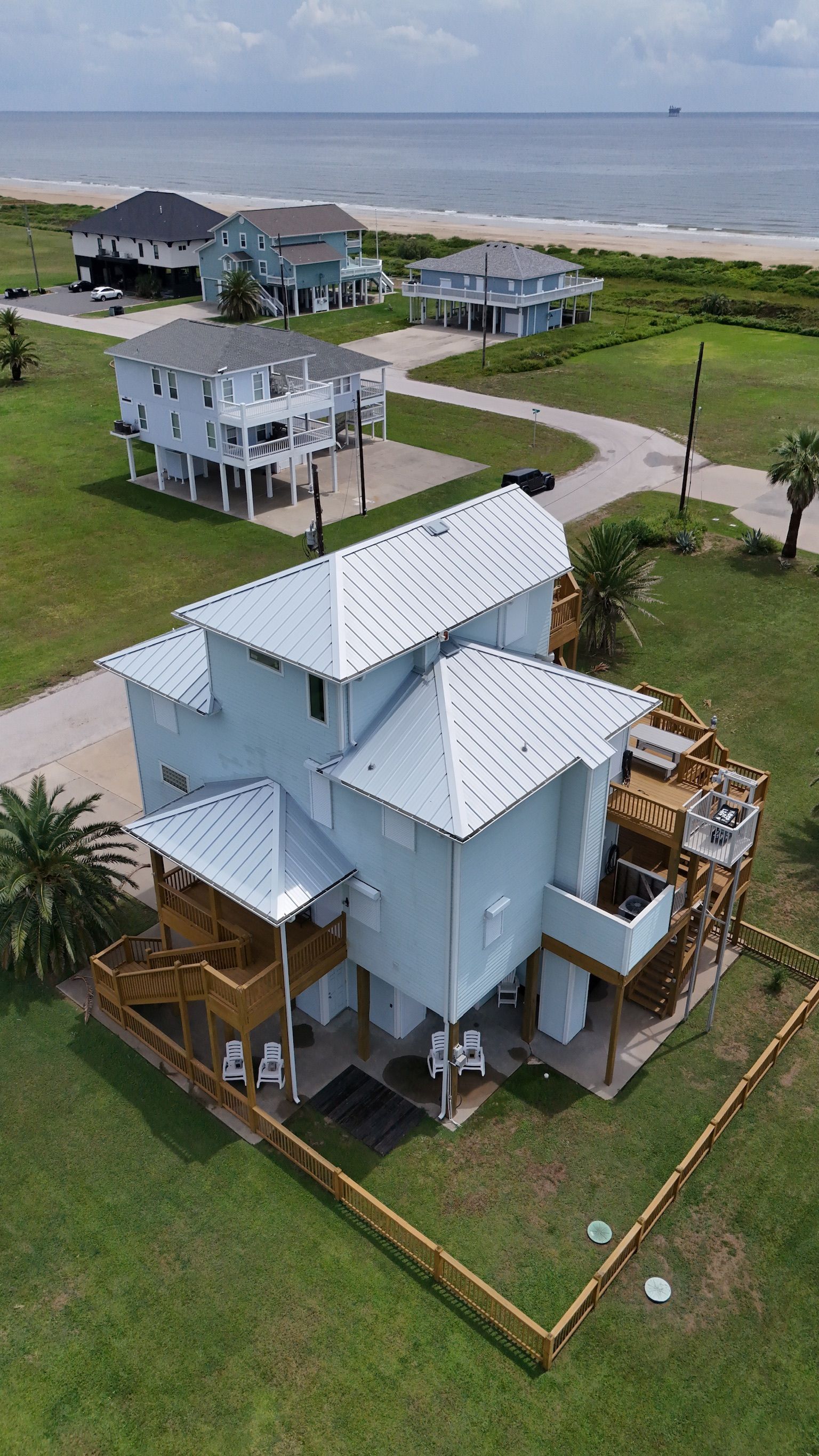Aerial view of several blue beach houses with a green lawn and ocean in the background.