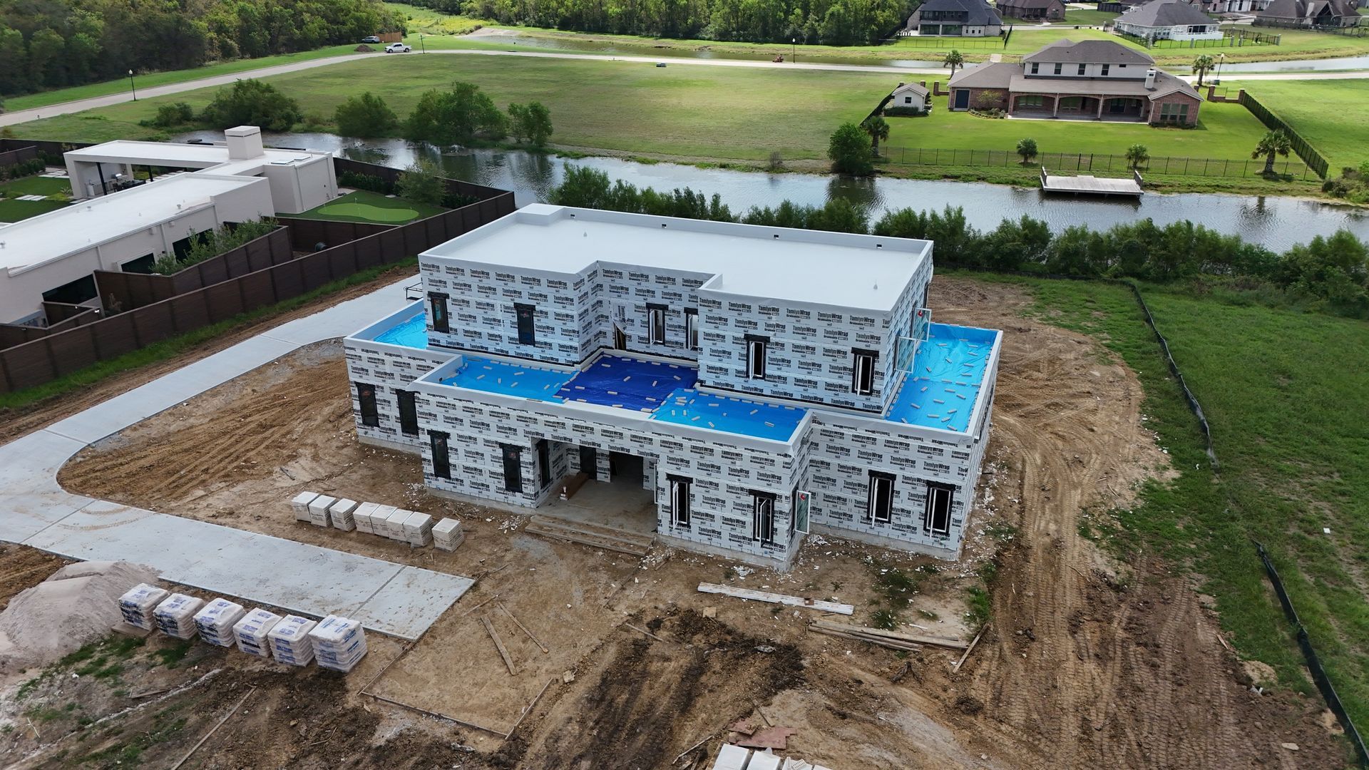 House under construction, light-colored walls covered in wrapping, blue patches on roof, surrounded by dirt, water, and grass.