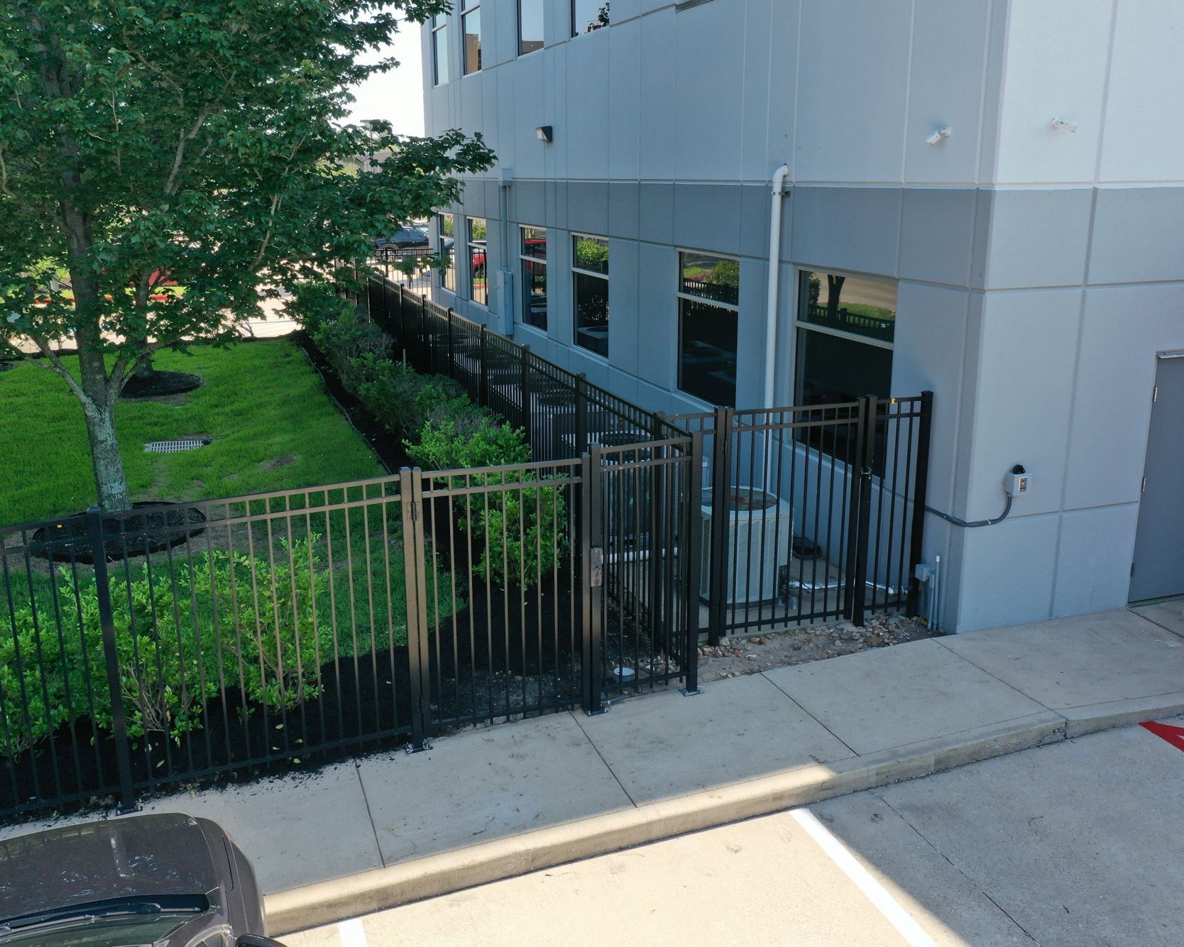 Black metal fence enclosing an AC unit next to a gray building and a grassy area with a tree.