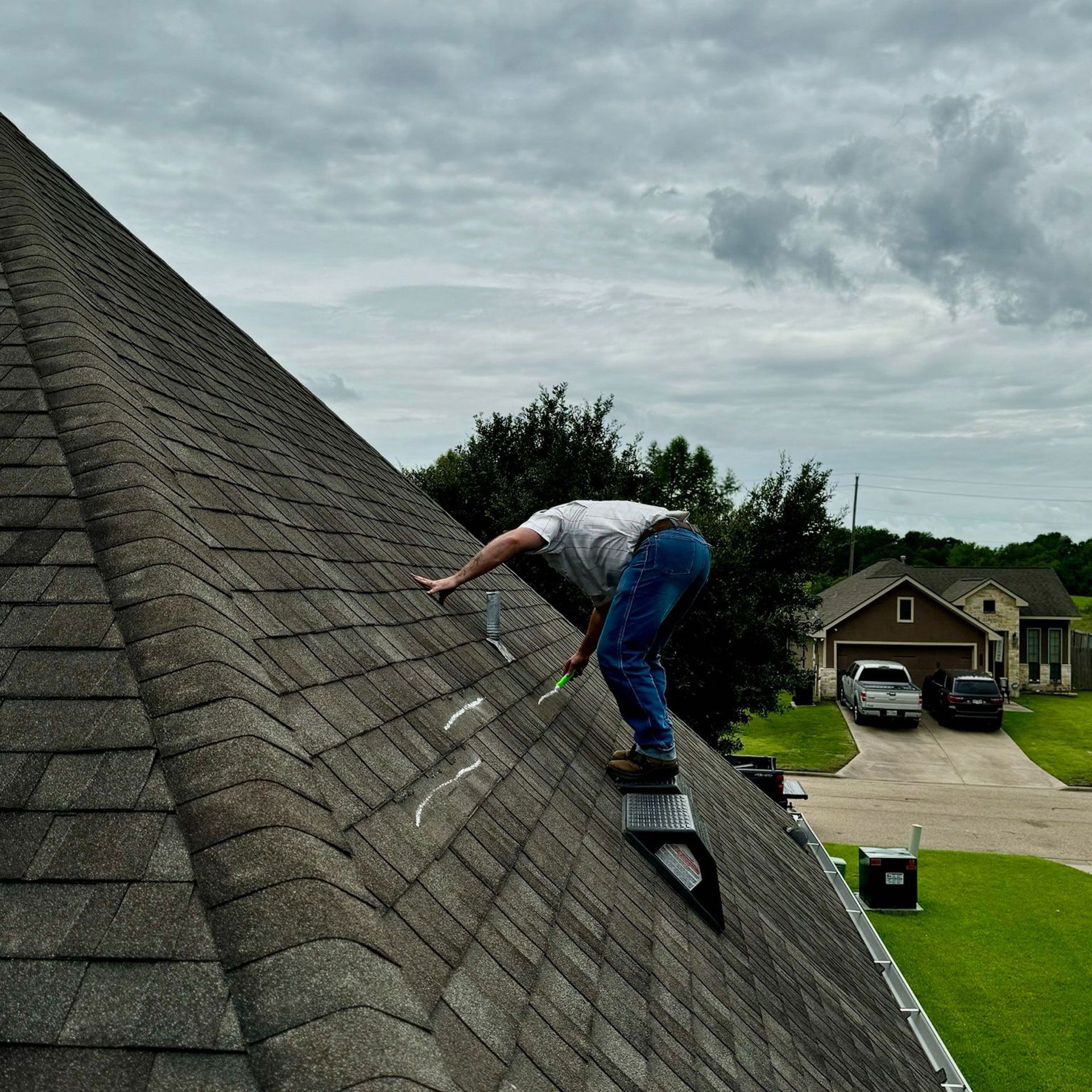 Person on a steeply sloped roof with tools, reaching toward the top of the roof. Cloudy sky, residential area.