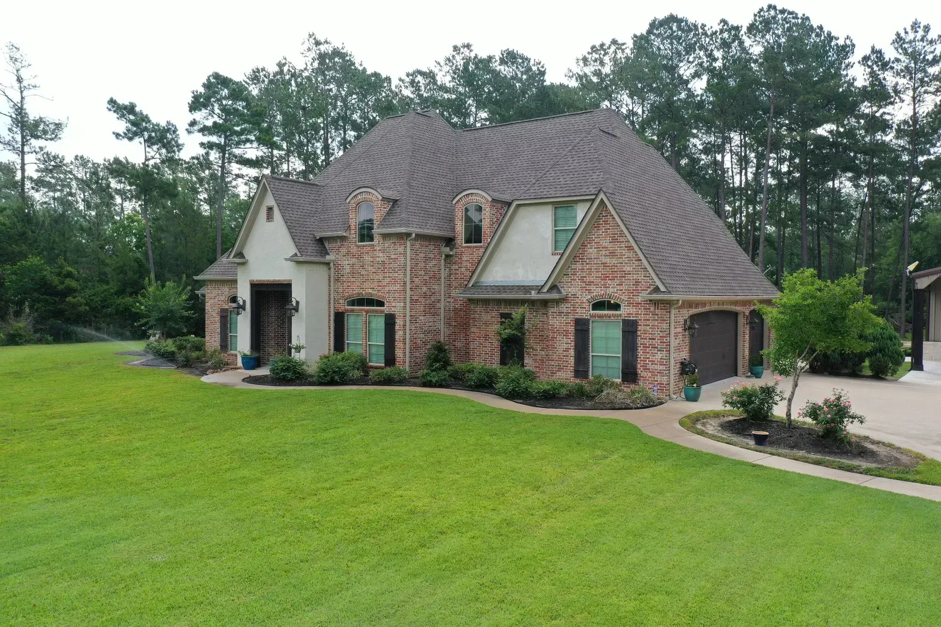 Two-story brick house with a brown roof and shutters, a green lawn, and trees in the background.