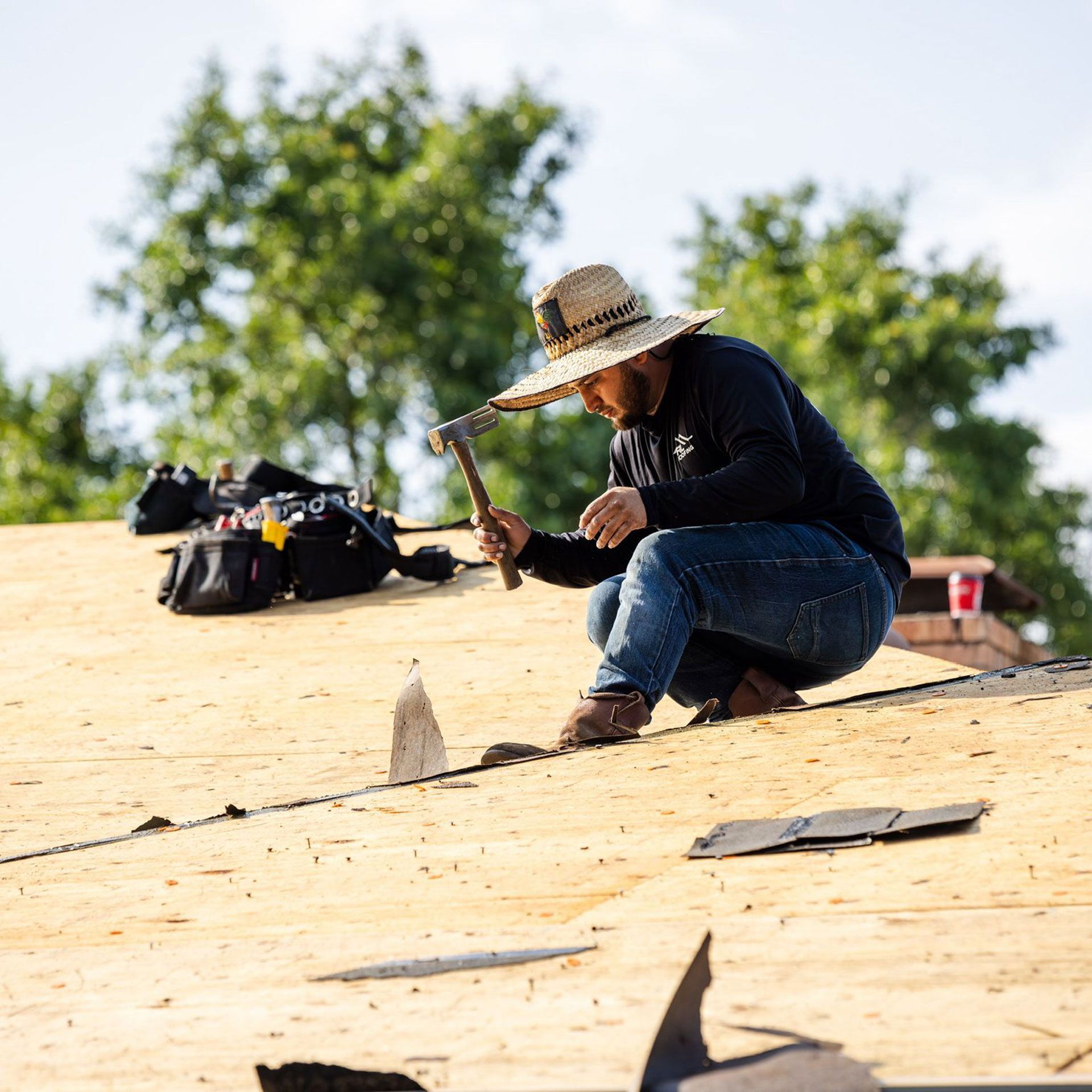 Roofer wearing straw hat hammers shingles on a rooftop. Tool bag and trees in background.