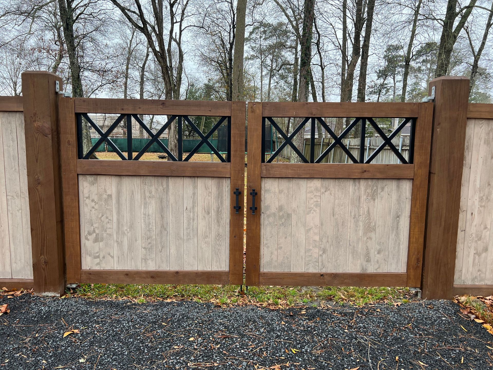 Wooden gate with dark cross design, matching fence, brown posts, gravel ground, and trees in the background.