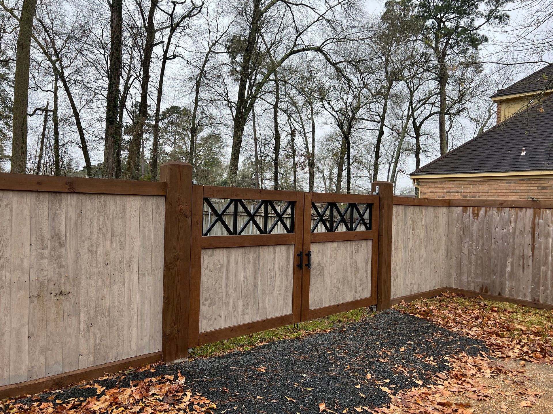 Wooden fence and gate with decorative black cross-hatching, gravel path, and fall leaves.