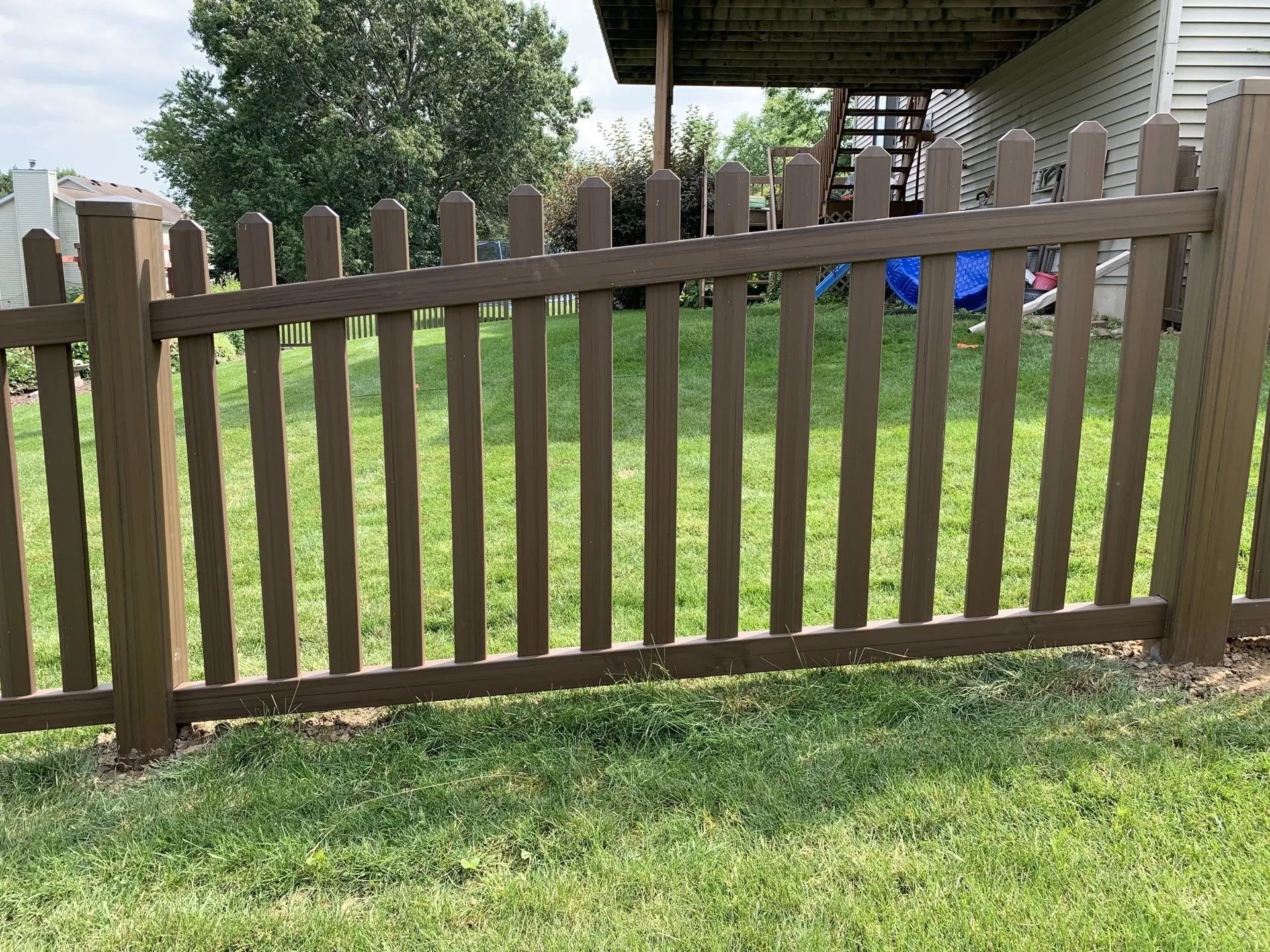 Brown picket fence in a grassy yard, partially obscuring a backyard and house.