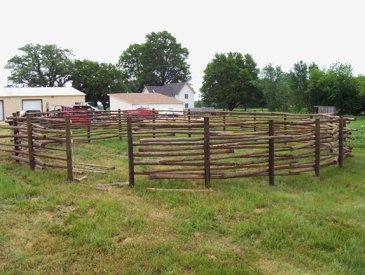 Round wooden corral in a grassy field, with buildings and trees in the background under an overcast sky.