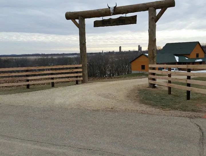 Entrance to a ranch with wooden gate, sign, and fence. A building with a green roof is visible. Cloudy sky.