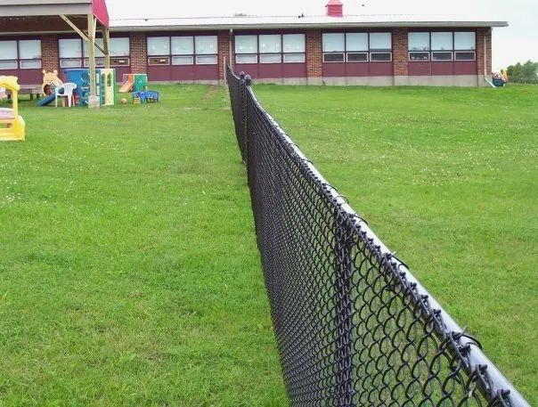 Black chain-link fence in front of a green grassy field with a brick building in the background.