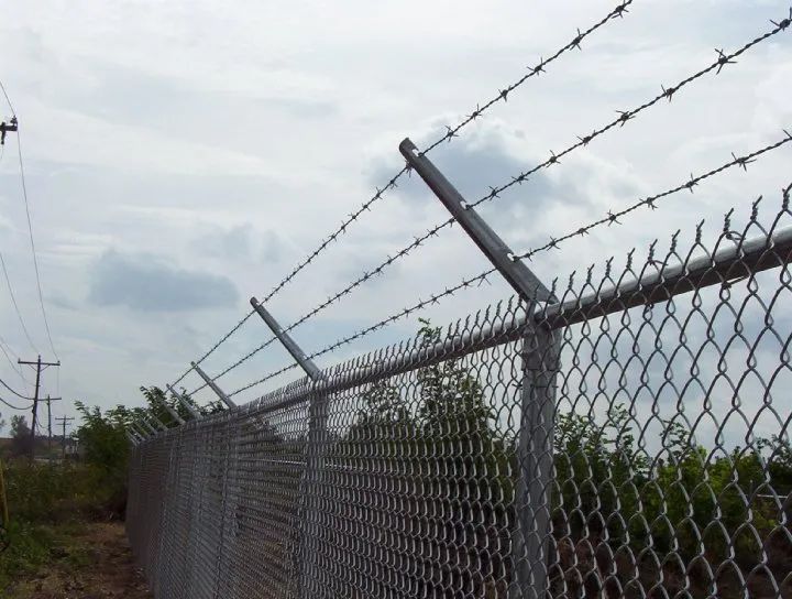 Chain-link fence topped with barbed wire, outdoors under a cloudy sky.