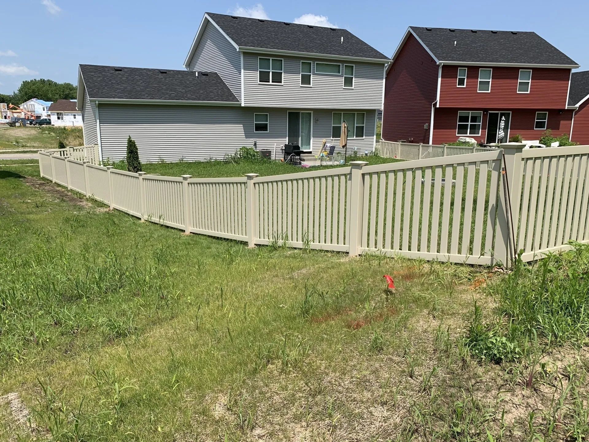 Beige fence encloses a backyard with a two-story gray and red house. Green grass in foreground.