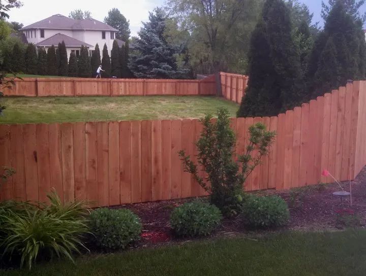 Wooden fence encloses a green lawn, garden with bushes and trees, a house in the background.