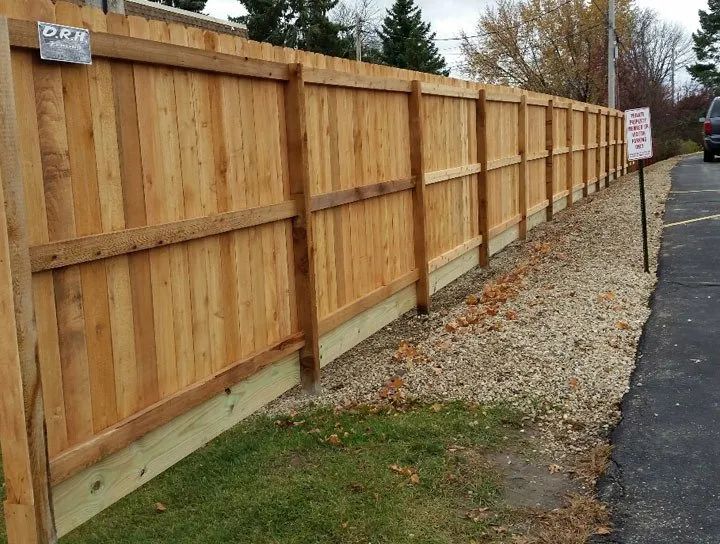 Wooden fence next to a gravel bed and asphalt. Brown boards and posts with a grassy edge.