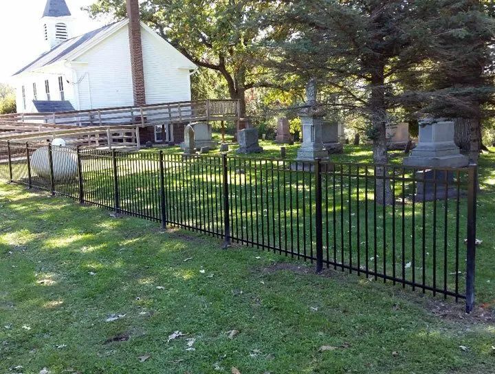 Black metal fence in a green lawn surrounds a cemetery with headstones and a white building with a ramp.