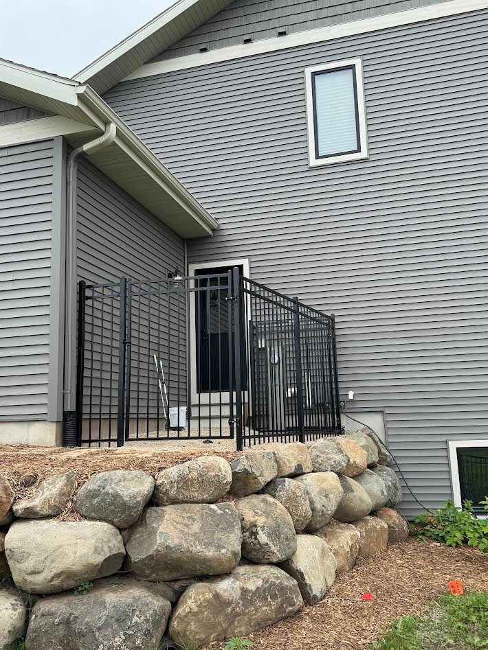 Black metal railing surrounds a small patio area by a gray house with a stone retaining wall.
