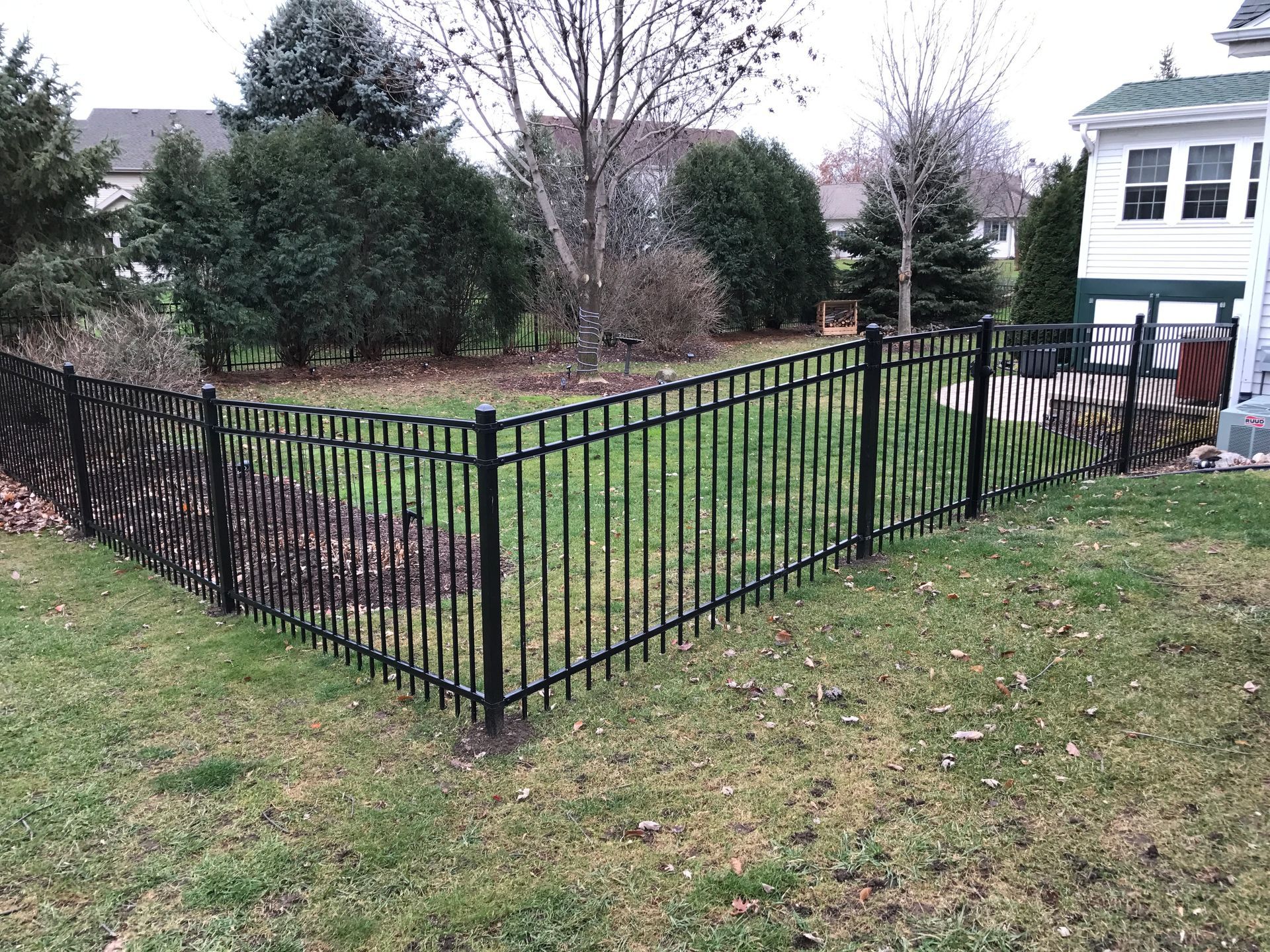 Black metal fence enclosing a grassy yard, with trees and a house in the background.