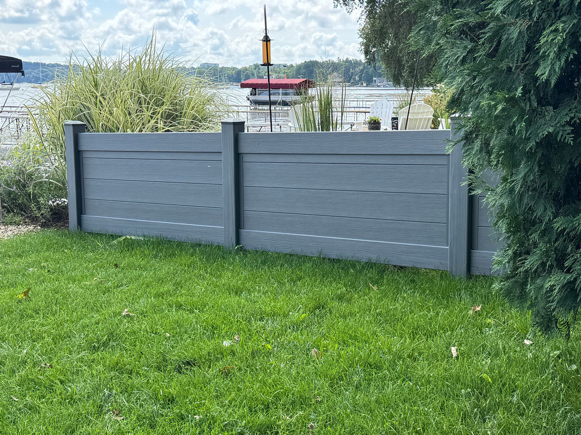 Gray composite fence with green lawn in front and lake in background.
