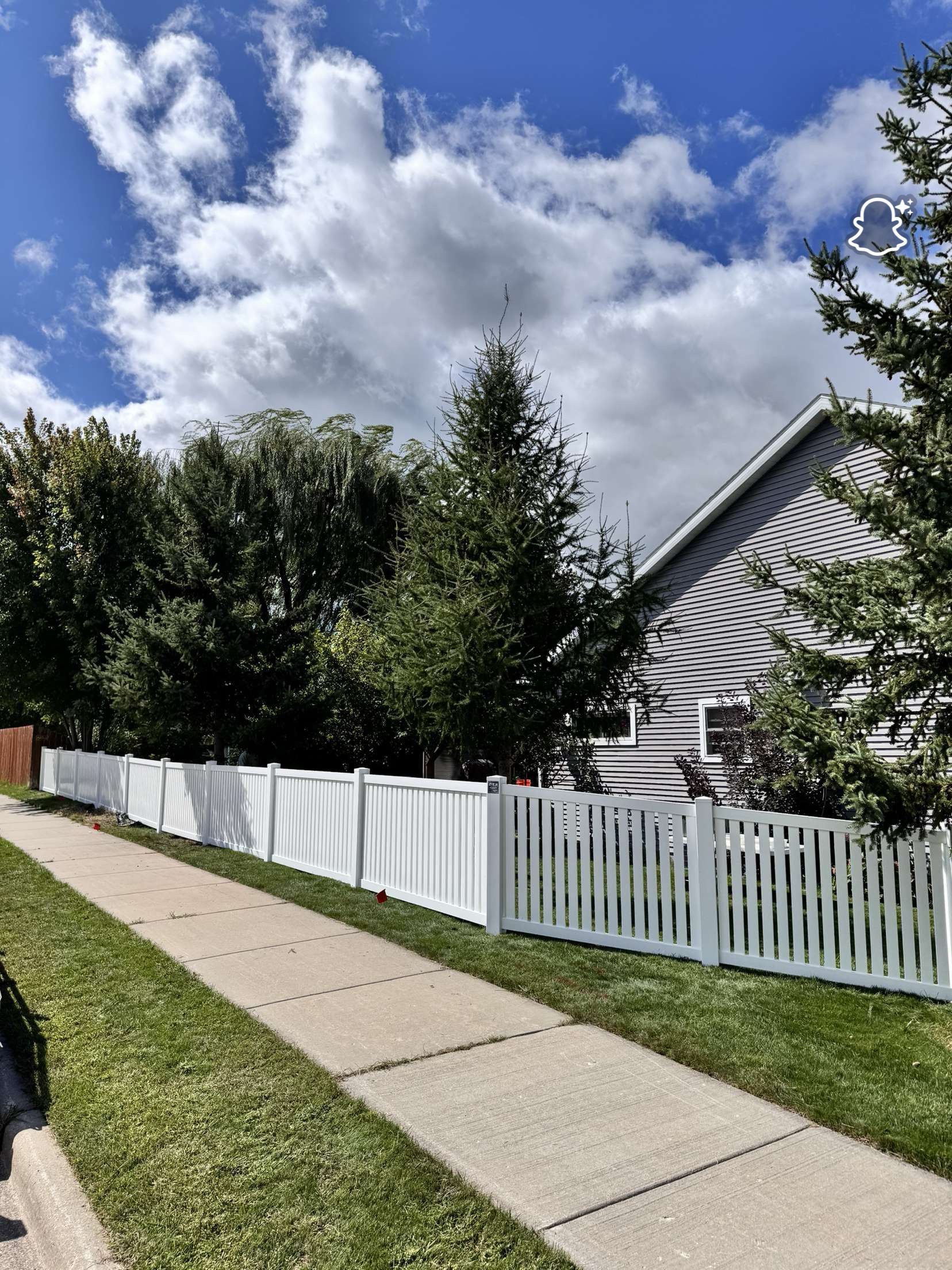 White picket fence along a sidewalk, with trees and a house in the background under a cloudy sky.