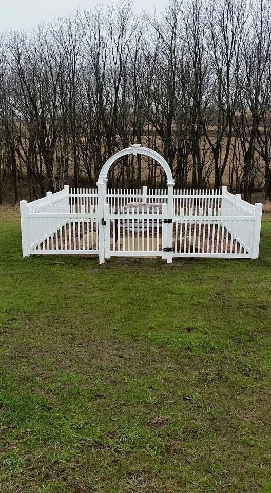 White picket fence with arched gate in a grassy field, trees in the background.