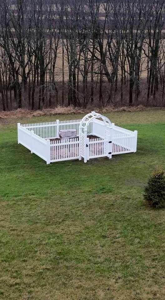 White picket fence encloses an outdoor space with an arched entrance, set on a green lawn with a treeline background.