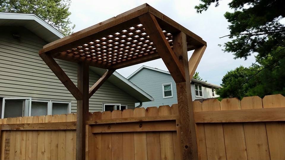 Wooden arbor with lattice roof over a wooden fence, in a yard with houses in background.