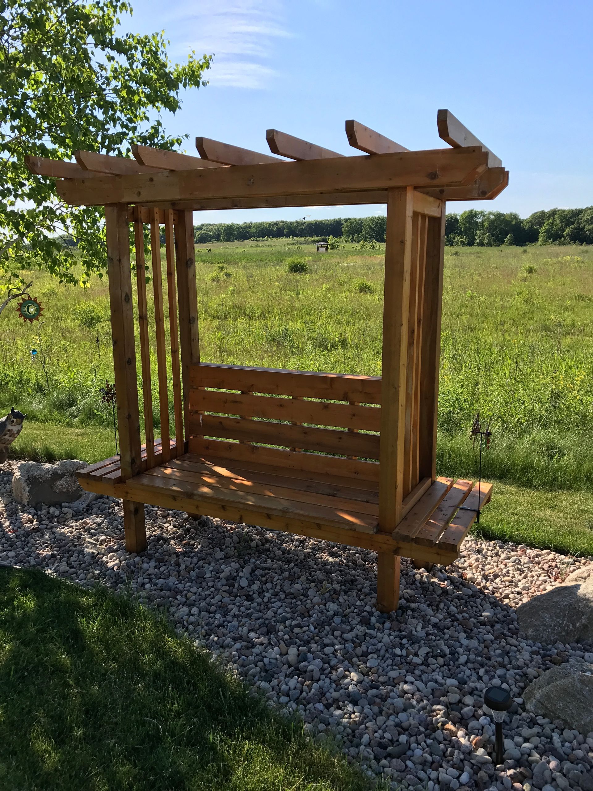 Wooden bench with overhead pergola in a field.