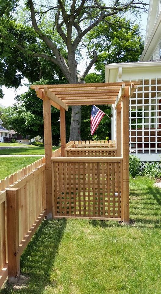 Wooden fence with pergola structure, American flag, and house in background. Green grass.