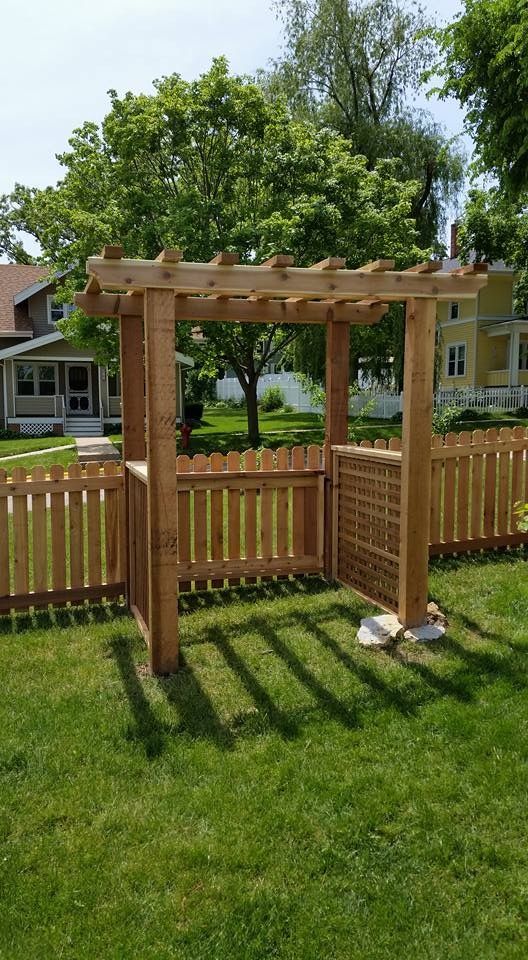 Wooden pergola with fence, set in a grassy yard, with a light brown color. Houses and trees in the background.