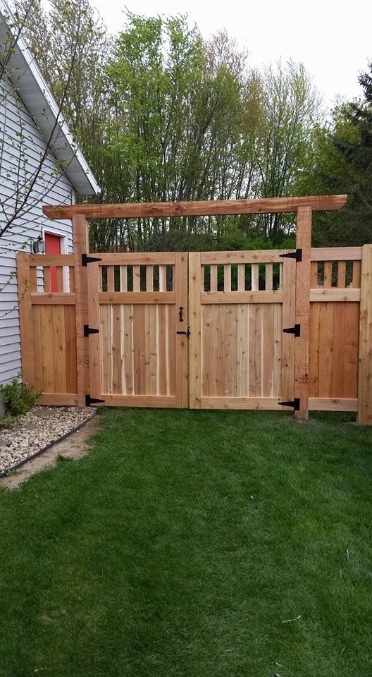 Wooden gate with overhead beam, leading to a yard with green grass, surrounded by wooden fence.