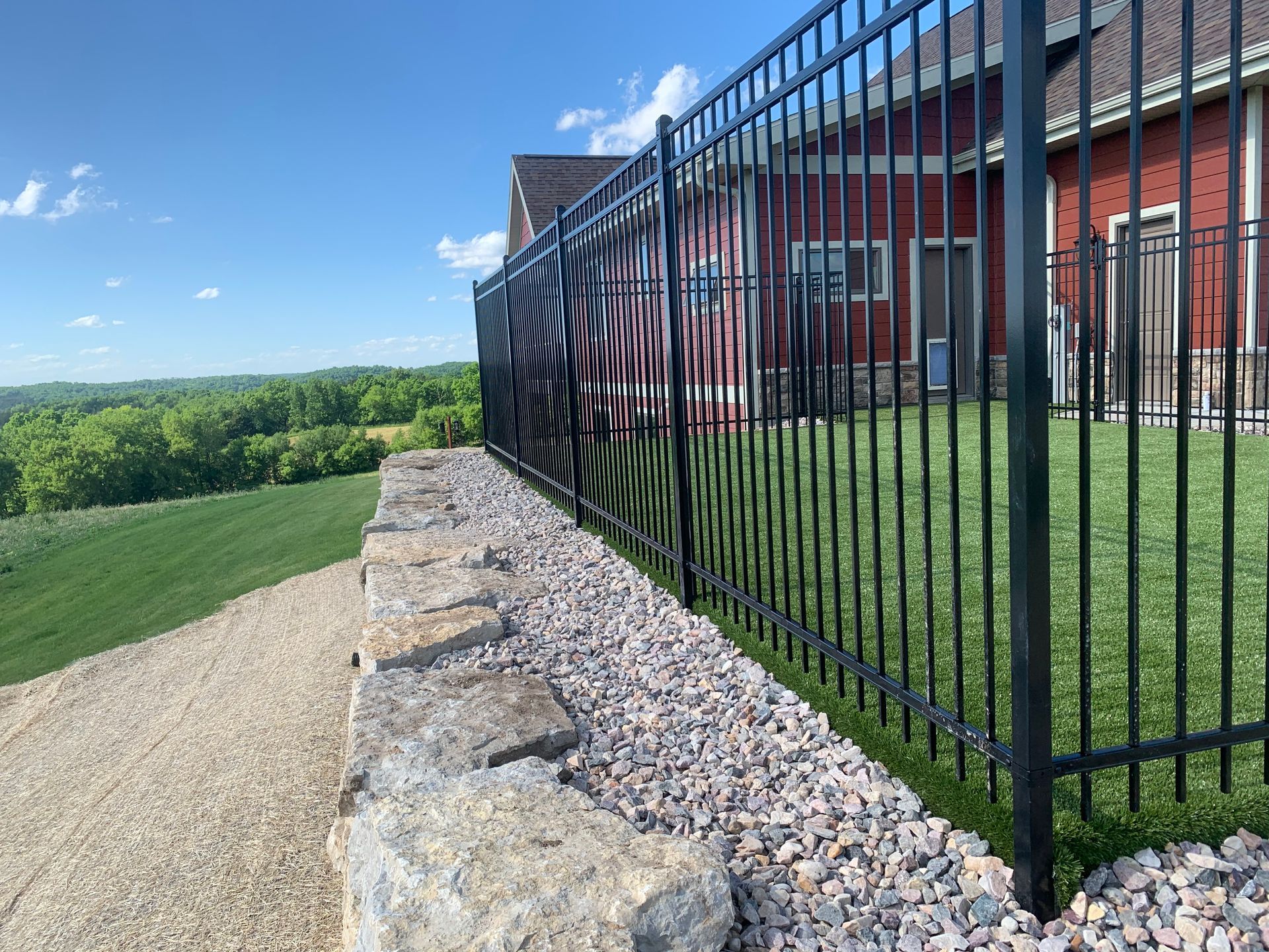 Black metal fence atop a stone wall, bordering a gravel path and green lawn with a red building in the background.