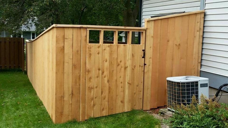 Wooden fence enclosing an AC unit next to a house with a small square window near the top.